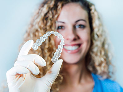 A woman holds up a clear dental retainer with her left hand, smiling and looking directly at the camera.