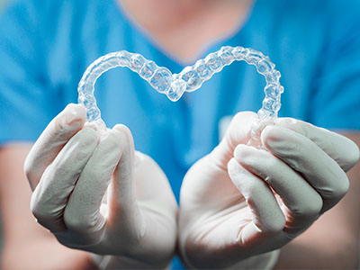 The image shows a pair of clear braces held by a person wearing white gloves and a blue surgical gown, with a heart shape formed by the braces.