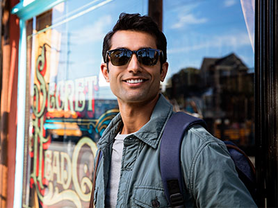 The image features a man standing outdoors in front of a storefront window with sunglasses on his face, wearing a backpack and a light-colored jacket, posing for the camera.